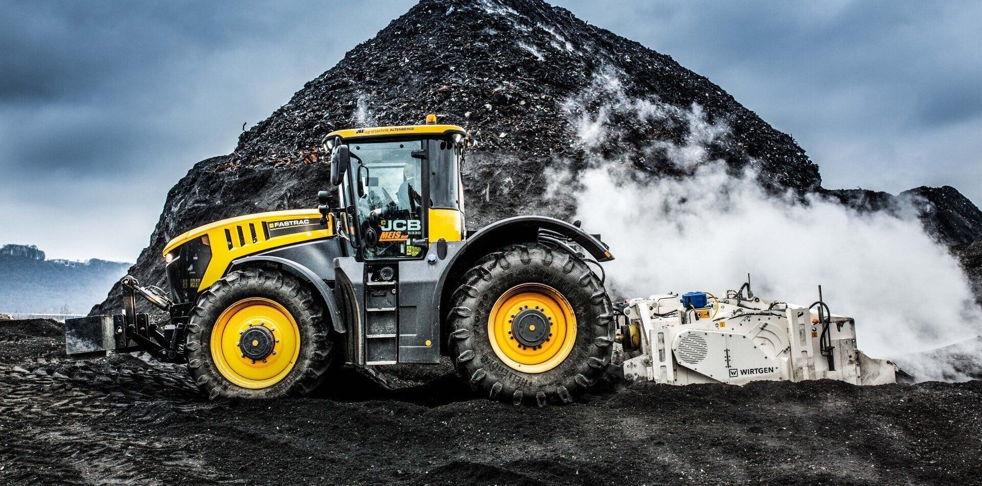 Ein gelb-schwarzer JCB-Traktor fährt in der Nähe eines großen dunklen Erdhügels und stößt Dampf aus einem weißen Maschinenaufsatz aus.
