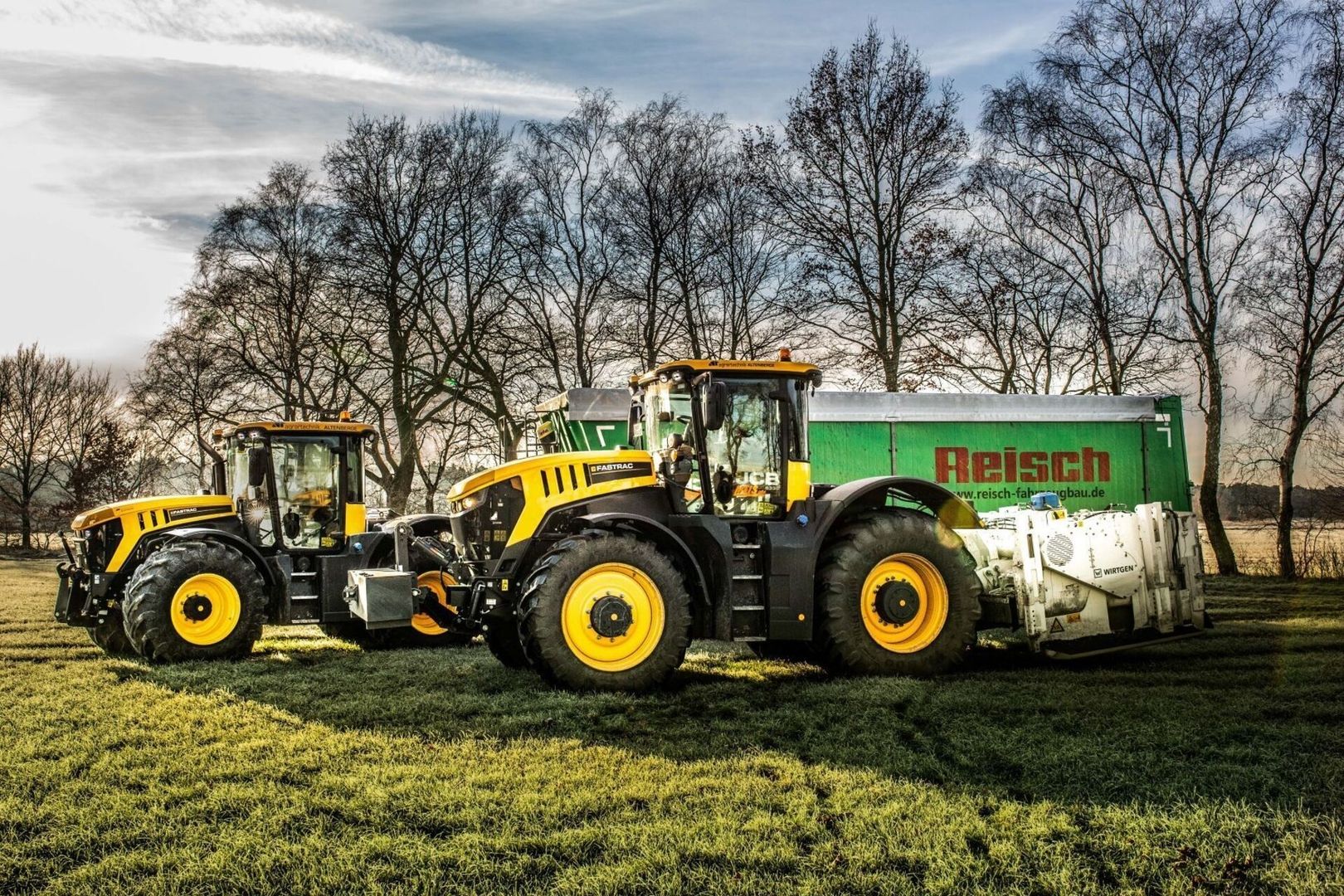 Zwei gelb-schwarze JCB-Traktoren, die auf einem Feld geparkt sind, mit einem grünen Reisch-Anhänger dahinter und kahlen Bäumen im Hintergrund.