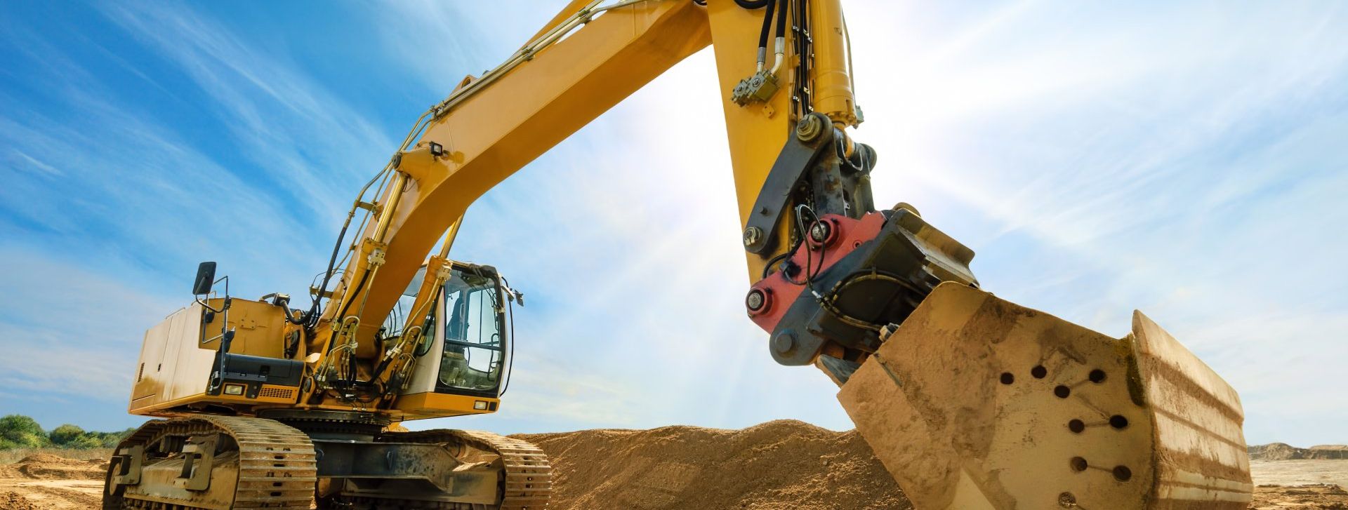 Ein großer gelber Bagger mit erhobenem Arm und Schaufel steht auf einer Baustelle unter blauem Himmel.